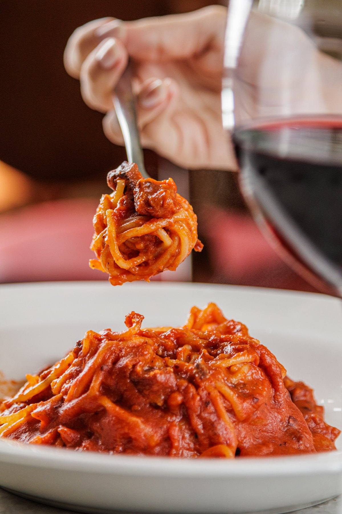 Garfo erguendo spaghetti all’amatriciana, com taça de vinho tinto ao lado